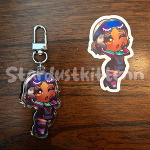 May include: Two items: a keychain and a sticker. Both feature a cartoon character with brown skin, wearing a purple and black outfit with green accents. The keychain has a silver clasp. The sticker is a die-cut shape.