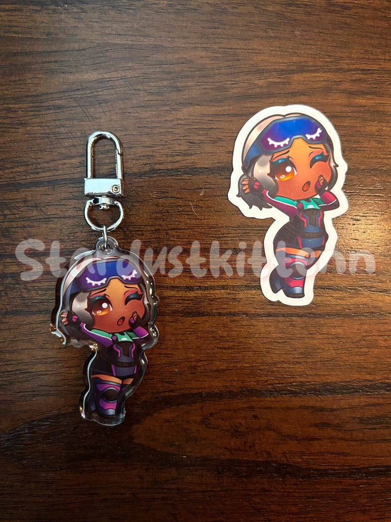 May include: Two items: a keychain and a sticker. Both feature a cartoon character with brown skin, wearing a purple and black outfit with green accents. The keychain has a silver clasp. The sticker is a die-cut shape.