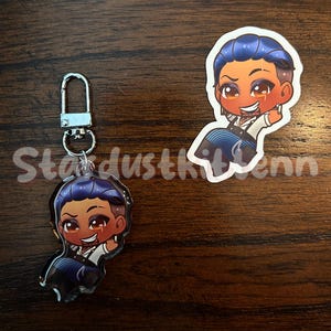 May include: Two items: a keychain charm and a sticker. Both feature a cartoon character with blue hair, a white shirt, and a black vest. The keychain has a silver clasp. The sticker is a die-cut shape. The text "Stardustkitten" is partially visible.