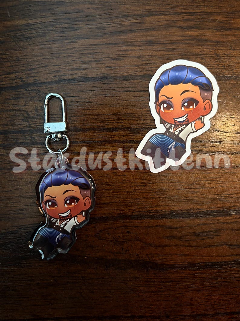 May include: Two items: a keychain charm and a sticker. Both feature a cartoon character with blue hair, a white shirt, and a black vest. The keychain has a silver clasp. The sticker is a die-cut shape. The text "Stardustkitten" is partially visible.