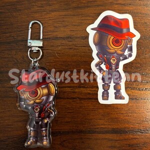 May include: A keychain and sticker featuring a robot character with a red hat, gold accents, and a mustache. The keychain has a silver clasp. The sticker is a die-cut shape of the same character.