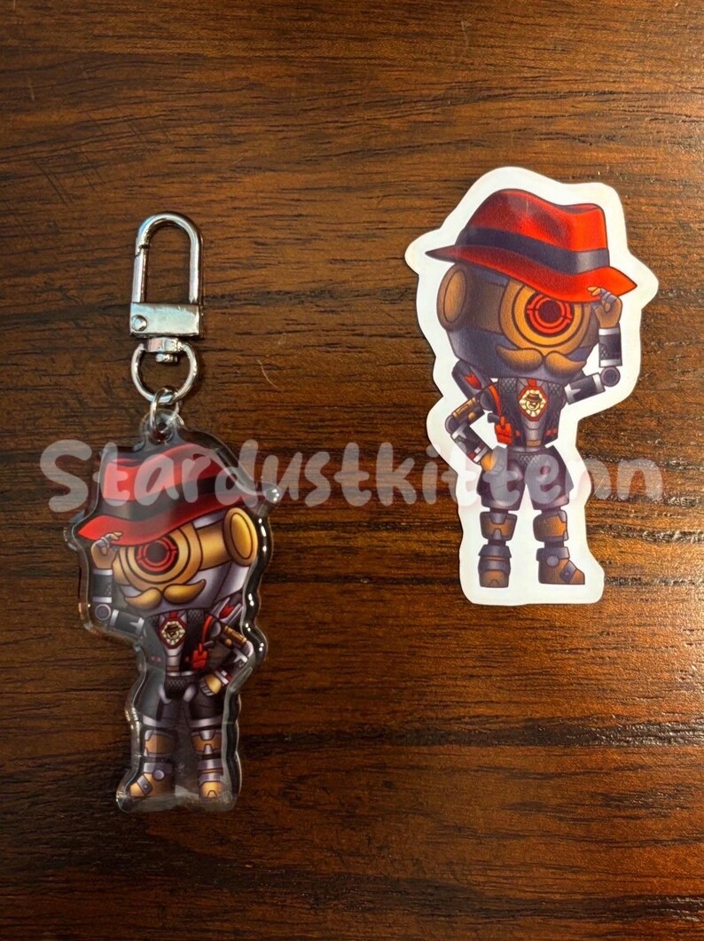 May include: A keychain and sticker featuring a robot character with a red hat, gold accents, and a mustache. The keychain has a silver clasp. The sticker is a die-cut shape of the same character.