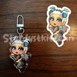 May include: Two items: a clear acrylic charm with a silver clasp and a sticker. Both feature a cartoon character with pale skin, gray hair, and blue accents. The character wears a black outfit with a skull design and holds a small skull. The charm has the text "Skydustkitten".