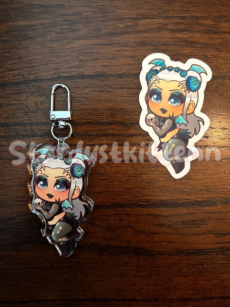 May include: Two items: a clear acrylic charm with a silver clasp and a sticker. Both feature a cartoon character with pale skin, gray hair, and blue accents. The character wears a black outfit with a skull design and holds a small skull. The charm has the text "Skydustkitten".