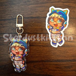 May include: An acrylic keychain and sticker featuring a cartoon character with blonde hair, glasses, and a blue hat. The character is wearing a colorful outfit and holding a phone. The keychain has a gold clasp.