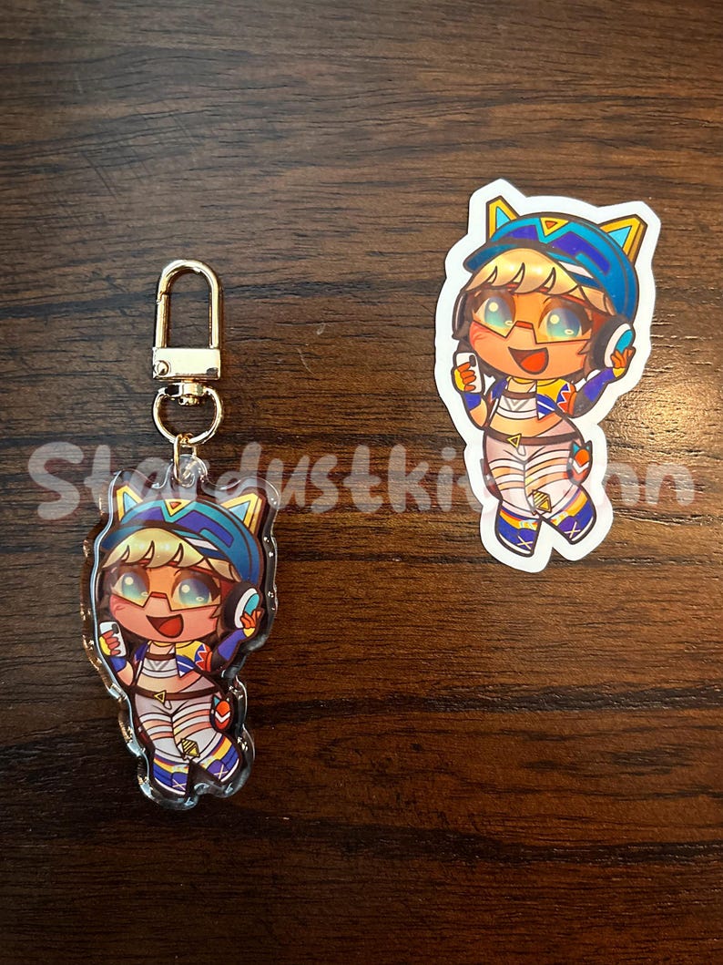May include: An acrylic keychain and sticker featuring a cartoon character with blonde hair, glasses, and a blue hat. The character is wearing a colorful outfit and holding a phone. The keychain has a gold clasp.