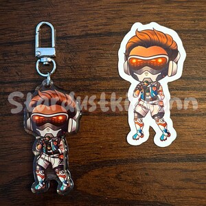 May include: Two items: a keychain and a sticker featuring a cartoon character with orange hair, a futuristic helmet, and a white and blue outfit. The keychain has a silver clasp. The sticker is a die-cut shape.