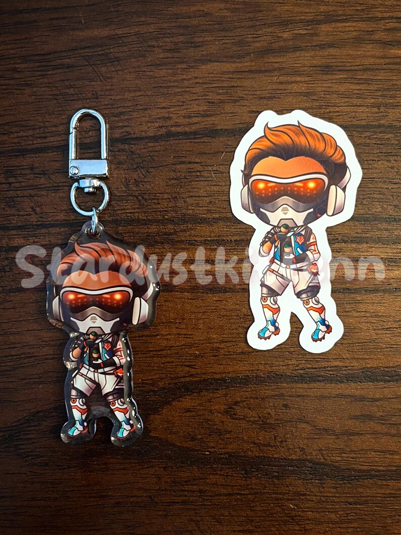 May include: Two items: a keychain and a sticker featuring a cartoon character with orange hair, a futuristic helmet, and a white and blue outfit. The keychain has a silver clasp. The sticker is a die-cut shape.