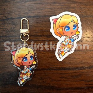 Op de afbeelding: Een heldere acryl sleutelhanger en sticker met een stripfiguur met blond haar, blauwe ogen en een ster. Het personage draagt een kleurrijke outfit. De sleutelhanger heeft een goudkleurige sluiting. De tekst "Stardustkinn" is zichtbaar.