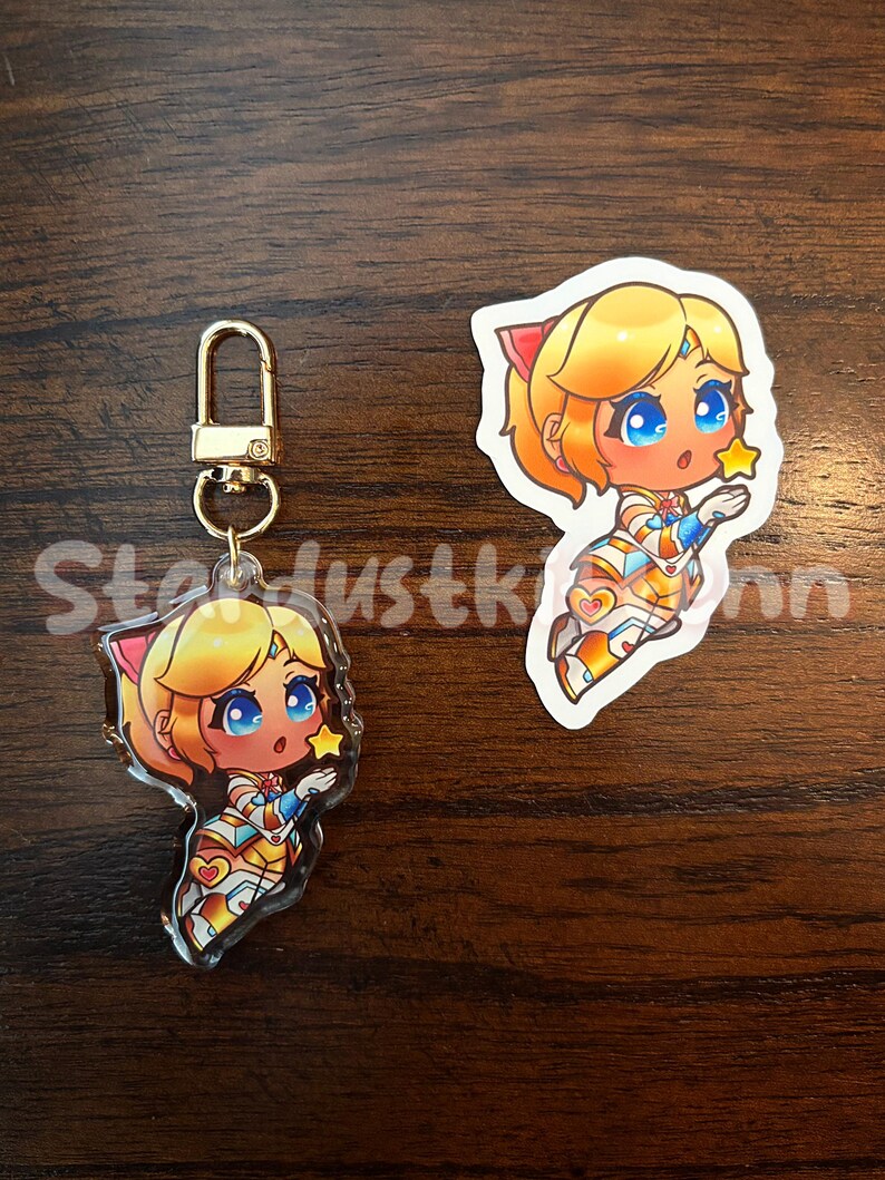 Op de afbeelding: Een heldere acryl sleutelhanger en sticker met een stripfiguur met blond haar, blauwe ogen en een ster. Het personage draagt een kleurrijke outfit. De sleutelhanger heeft een goudkleurige sluiting. De tekst "Stardustkinn" is zichtbaar.