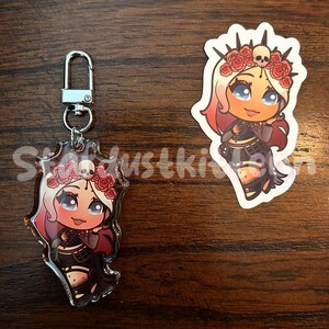 May include: A keychain and sticker featuring a cartoon character with white and red hair, a floral crown, and a black outfit. The keychain has a silver clasp. The sticker is white with a die-cut shape.