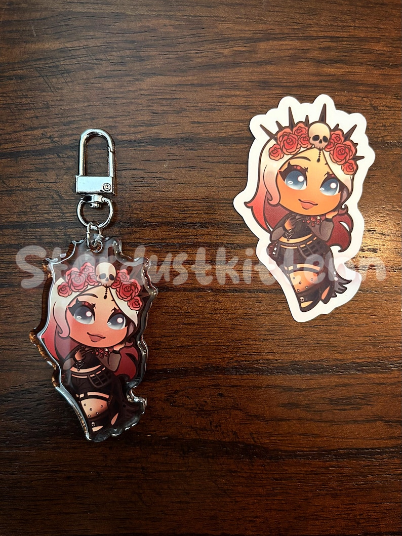 May include: A keychain and sticker featuring a cartoon character with white and red hair, a floral crown, and a black outfit. The keychain has a silver clasp. The sticker is white with a die-cut shape.