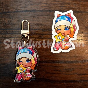 Op de afbeelding: Een heldere acryl sleutelhanger en sticker met een stripfiguur met blauwe ogen, oranje haar en een ster. Het personage draagt een wit en blauw hoedje en een roze outfit. De sleutelhanger heeft een goudkleurige sluiting.