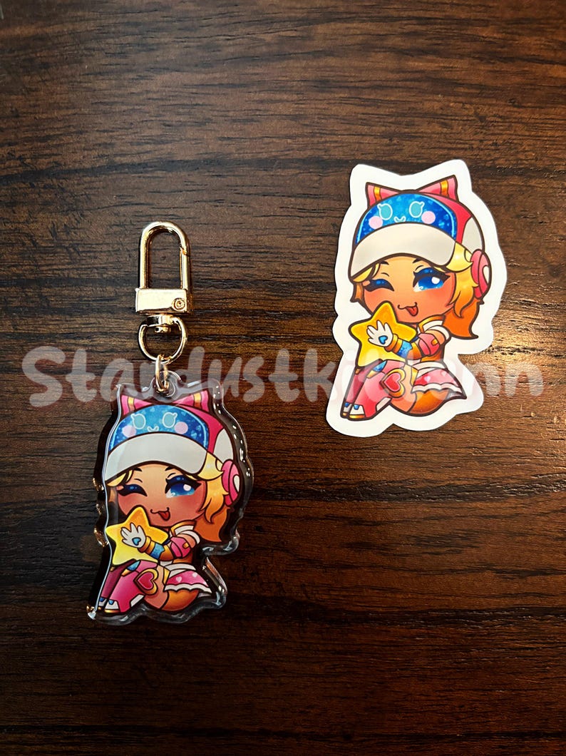 Op de afbeelding: Een heldere acryl sleutelhanger en sticker met een stripfiguur met blauwe ogen, oranje haar en een ster. Het personage draagt een wit en blauw hoedje en een roze outfit. De sleutelhanger heeft een goudkleurige sluiting.