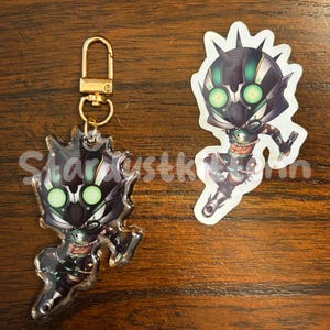 May include: A clear acrylic keychain and sticker featuring a stylized character with a black and green helmet and glowing green eyes. The keychain has a gold-colored clasp. The sticker is white with a jagged edge.