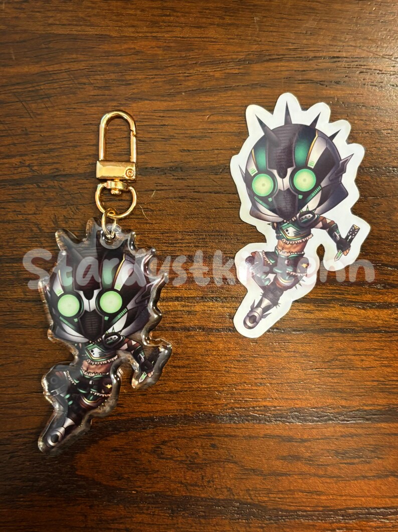 May include: A clear acrylic keychain and sticker featuring a stylized character with a black and green helmet and glowing green eyes. The keychain has a gold-colored clasp. The sticker is white with a jagged edge.