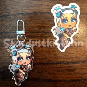 May include: Two items: a keychain and a sticker featuring a cartoon character with blue and white hair, blue accents, and a dark outfit. The keychain has a silver clasp. The sticker is a similar design. The background is a wooden surface.