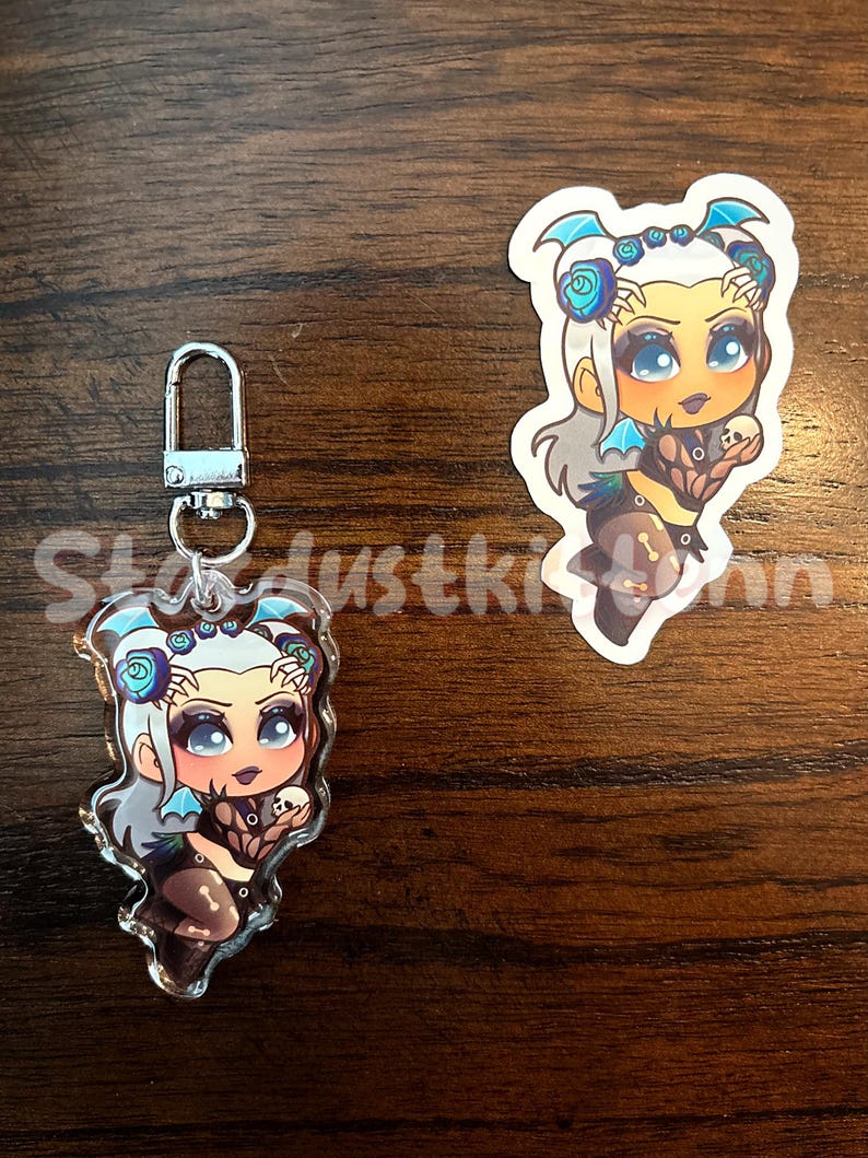 May include: Two items: a keychain and a sticker featuring a cartoon character with blue and white hair, blue accents, and a dark outfit. The keychain has a silver clasp. The sticker is a similar design. The background is a wooden surface.