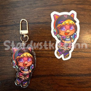 May include: A clear acrylic keychain and sticker featuring a cartoon character with pink hair, glasses, and headphones. The character is wearing a colorful outfit with black, pink, and purple accents. The keychain has a gold-colored clasp.