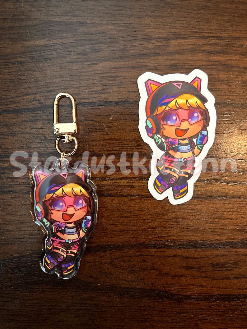 May include: A clear acrylic keychain and sticker featuring a cartoon character with pink hair, glasses, and headphones. The character is wearing a colorful outfit with black, pink, and purple accents. The keychain has a gold-colored clasp.