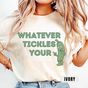 Woman wearing an ivory Comfort Colors T-shirt featuring the phrase “Whatever Tickles Your Pickle” in green retro lettering with a cute illustrated pickle character beside the text. Casual unisex graphic tee shown styled with light denim for a fun pickle-themed shirt design.