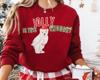 May include: A cardinal red sweatshirt with the text "JOLLY IS THIS ENOUGH?" and a graphic of a polar bear wearing a Santa hat. The sweatshirt is part of a Christmas-themed collection and is also available in a shirt and hoodie.