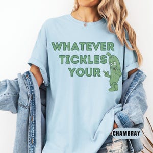 Woman wearing an chambray Comfort Colors T-shirt featuring the phrase “Whatever Tickles Your Pickle” in green retro lettering with a cute illustrated pickle character beside the text. Casual unisex graphic tee shown styled with light denim for a fun pickle-themed shirt design.