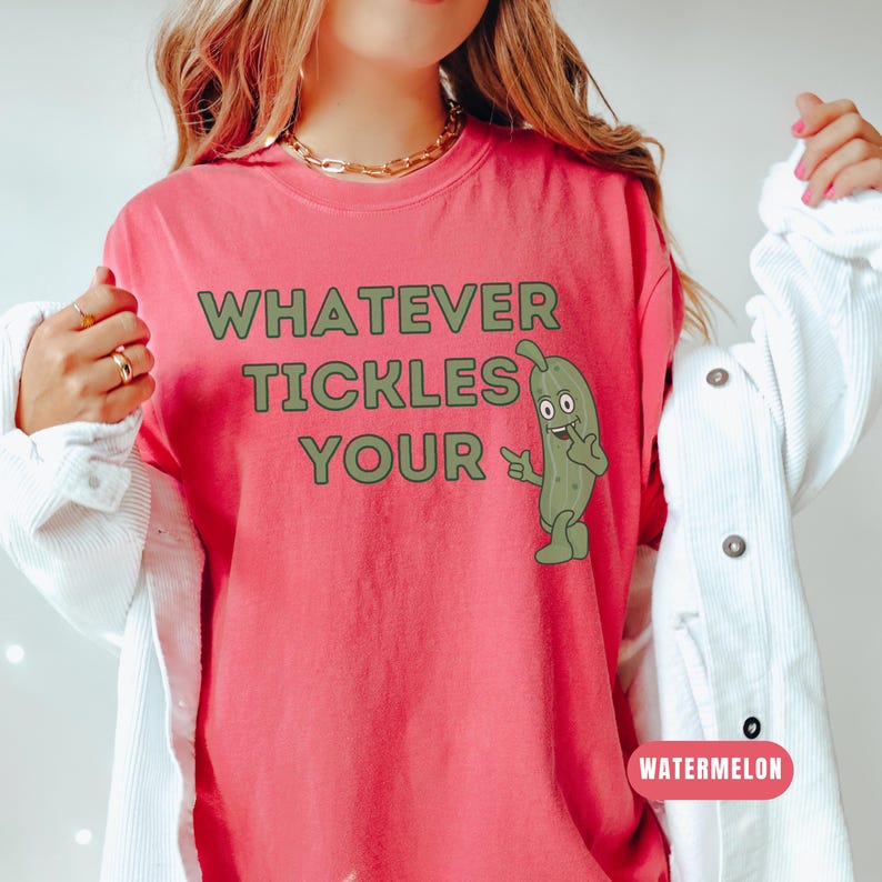 Woman wearing an watermelon Comfort Colors T-shirt featuring the phrase “Whatever Tickles Your Pickle” in green retro lettering with a cute illustrated pickle character beside the text. Casual unisex graphic tee shown styled with light denim for a fun pickle-themed shirt design.