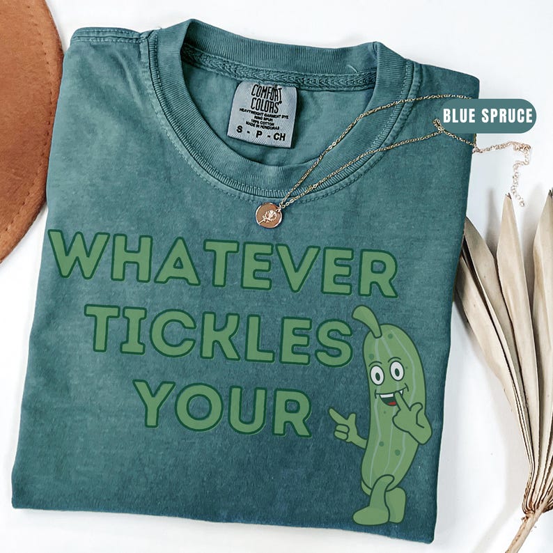Woman wearing an blue spruce Comfort Colors T-shirt featuring the phrase “Whatever Tickles Your Pickle” in green retro lettering with a cute illustrated pickle character beside the text. Casual unisex graphic tee shown styled with light denim for a fun pickle-themed shirt design.
