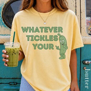 Woman wearing an Butter Comfort Colors T-shirt featuring the phrase “Whatever Tickles Your Pickle” in green retro lettering with a cute illustrated pickle character beside the text. Casual unisex graphic tee shown styled with light denim for a fun pickle-themed shirt design.