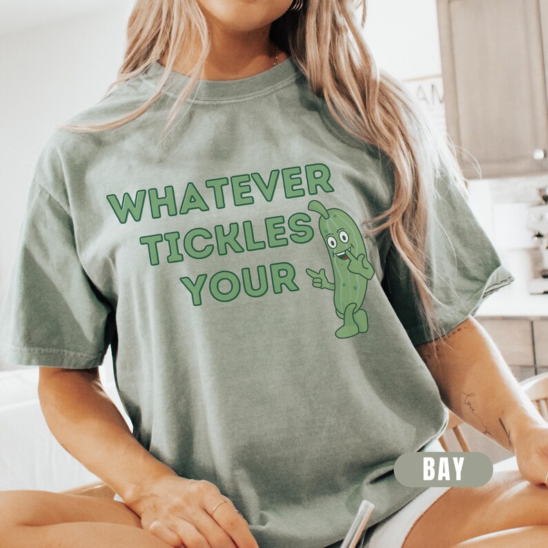 Woman wearing an bay Comfort Colors T-shirt featuring the phrase “Whatever Tickles Your Pickle” in green retro lettering with a cute illustrated pickle character beside the text. Casual unisex graphic tee shown styled with light denim for a fun pickle-themed shirt design.