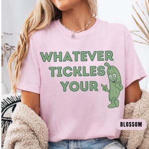 Woman wearing an blossom Comfort Colors T-shirt featuring the phrase “Whatever Tickles Your Pickle” in green retro lettering with a cute illustrated pickle character beside the text. Casual unisex graphic tee shown styled with light denim for a fun pickle-themed shirt design.