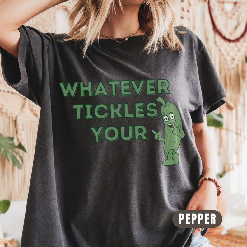 Woman wearing an pepper Comfort Colors T-shirt featuring the phrase “Whatever Tickles Your Pickle” in green retro lettering with a cute illustrated pickle character beside the text. Casual unisex graphic tee shown styled with light denim for a fun pickle-themed shirt design.
