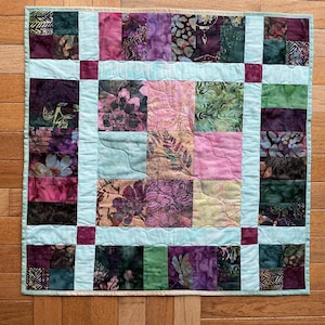 May include: A square quilt with a patchwork design. The quilt features squares of various colors, including pink, purple, green, and teal, with floral and abstract patterns. A teal border frames the quilt, with dark purple squares at the corners.