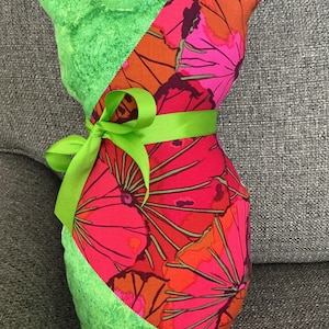 May include: A handmade cat-shaped pillow with a vibrant floral pattern in pink, orange, and purple. The cat's head and one side are a textured green fabric. A green ribbon is tied around the middle, creating a decorative accent.