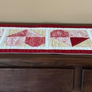 May include: A rectangular quilted table runner with a red border and a white inner border. The runner features four heart-shaped designs in red and cream fabrics, set against a light tan background. The runner is displayed on a dark wooden dresser.