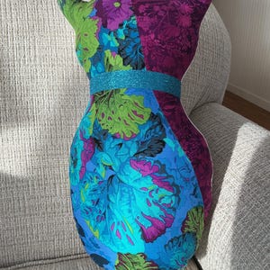 May include: A cat-shaped decorative pillow with a floral pattern in shades of blue, green, and purple. The pillow has a turquoise glitter band around the middle and pointed ears.