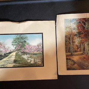 May include: Two framed landscape prints. One features a dirt road lined with pink flowering trees and a stone wall. The other depicts a forest scene with trees and a path. The prints are on aged paper.