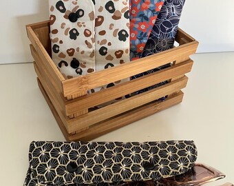 May include: A wooden crate holds several fabric eyeglass cases with various patterns, including a leopard print, floral, and geometric designs. A black and gold geometric patterned case with a pair of glasses is in front.
