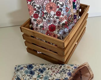 May include: A wooden crate containing several floral print spectacle cases in different colours. A matching case with blue floral print and brown sunglasses are in front. The cases have a button closure.