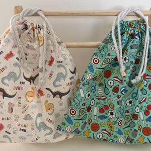 Puede incluir: Dos bolsas con cordón. Una es blanca con un estampado de dinosaurios y las palabras "Dino" y "Roar". La otra es de color turquesa con un estampado deportivo. Ambas bolsas tienen cordones blancos y cuelgan de un estante de madera.