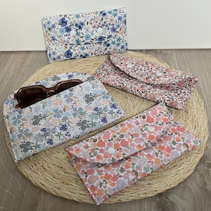 May include: A collection of floral print eyeglass cases in various colors, including blue, pink, and peach. One case is open, holding a pair of brown sunglasses. The cases are made of fabric and have a button closure. They are arranged on a woven mat.