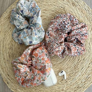 May include: Three floral scrunchies in pastel colors: blue, pink, and peach. The scrunchies are made of fabric with a small flower pattern. A white wireless earbud case and one earbud are also visible.