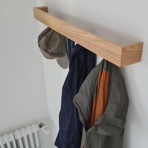Könnte beinhalten: Ein helles Holz-Kleiderregal an einer weißen Wand. Eine Baseballkappe, eine marineblaue Jacke und eine olivgrüne Jacke mit orangefarbenem Innenfutter hängen an dem Regal. Das Regal ist rechteckig und hat ein schlichtes, modernes Design.