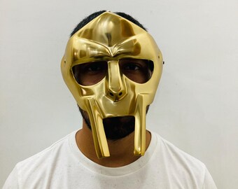 May include: A gold-colored mask with a distinctive, stylized design. The mask features a prominent nose, a wide mouth, and angular features. The person is wearing a white t-shirt with the text "US" and "DENIM COMPANY" printed on it.