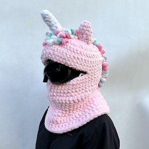 Könnte beinhalten: Rosa gestrickte Einhornmütze mit einem weißen Horn und einer flauschigen Mähne. Die Mütze hat eine schwarze Skimaske.