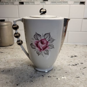 May include: White ceramic coffee pot with a silver lid and handle. The pot has a pink rose with gray leaves design.