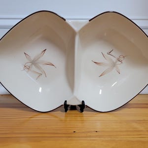 May include: Cream-colored, divided serving dish with a brown rim and leaf design. The dish has a unique shape with two compartments and is supported by a black stand. The leaf design features brown and gray tones.
