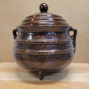 May include: A brown ceramic pot with a lid and three small feet. The pot has a textured surface and two handles on the sides. The lid has a small knob on top. The pot is likely used for cooking or storage.