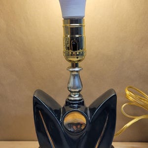 May include: A black and gold art deco style table lamp with a bright white LED bulb. The lamp has a unique, stylized base with gold accents and a gold-colored bulb socket. The lamp is on a wooden surface.